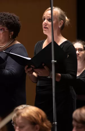 A White woman with straight dark hair, a Black woman with curly dark hair, and a White woman with blond hair in a bun sing in a chorus