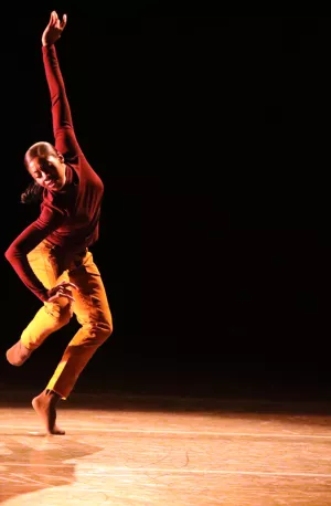 A Black woman in a burgundy shirt and tan pants dances on a stage with one arm extended in the air