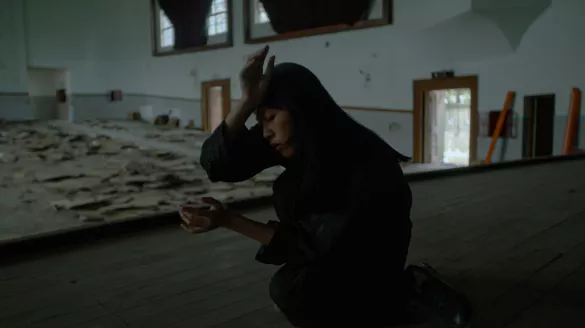 A woman with dark hair and clothing crouches with her palm at her forehead, with a large room filled with debris in the background