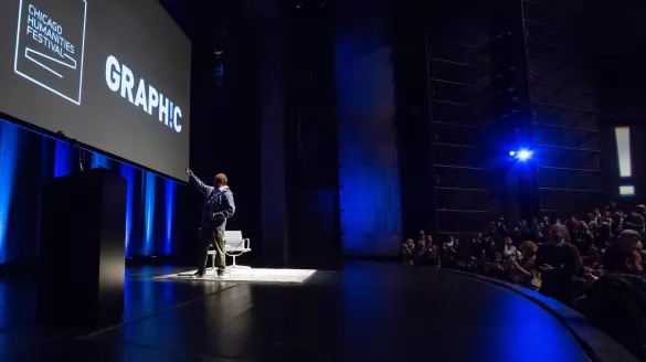 A man stands on a theater stage with blue accent lighting and a projected backdrop with white text that reads "Chicago Humanities Festival - GRAPHIC"