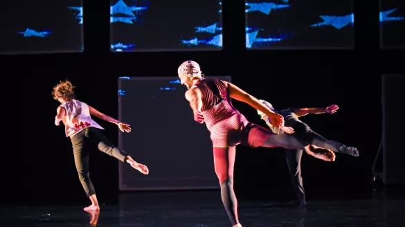 Three dancers in brightly colored clothing move across a a dark stage with blue cloud-like projections