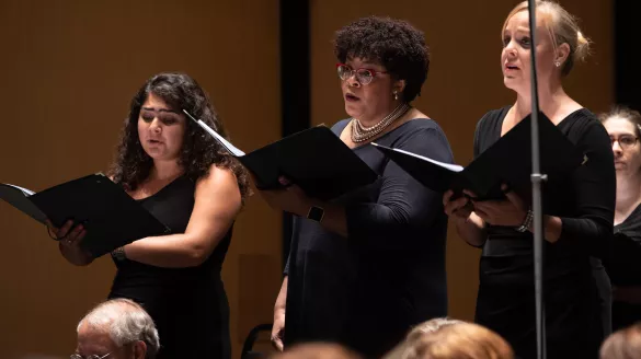 A White woman with straight dark hair, a Black woman with curly dark hair, and a White woman with blond hair in a bun sing in a chorus