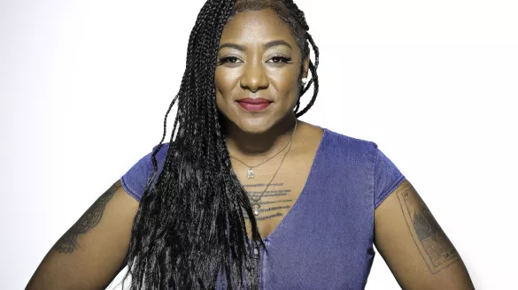 A Black woman with long braids and a blue shirt poses smiling, with her hands at her hips