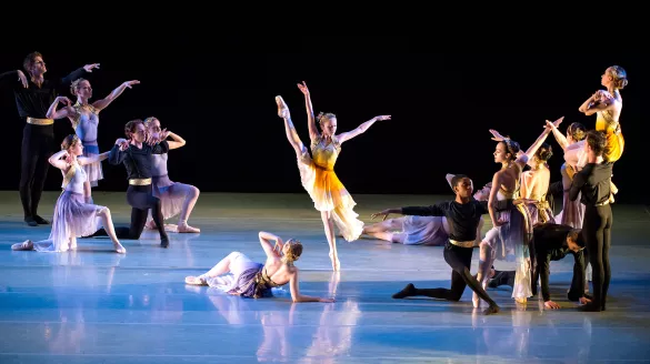 An ensemble of ballet dancers pose on a softly lit stage with blue hues, with a single female dancer at the center raising her leg high in the air