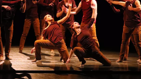 Dancers in brown sleeveless shirts and pants appear in standing and lunging positions on a black stage