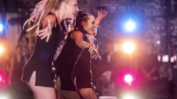 Close-up of three female dancers in black costumes, with blue, yellow, and red stage lights in the background