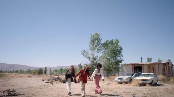 Three dancers moving their right foot forward in a desert-like town with two cars and a house