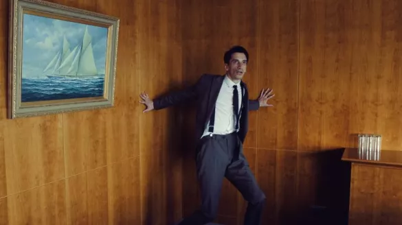 Man in suit leans against a wooden wall with a portrait of a sailboat on the left and a counter on the right.