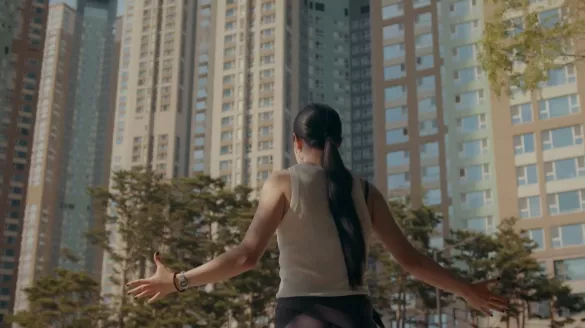 Woman wearing a dark black ponytail faces concrete buildings with her arms extended