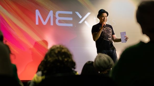 A man with a fedora hat stands in front of a crowd speaking into a microphone in front of a wall that projects the text "MEX talks"