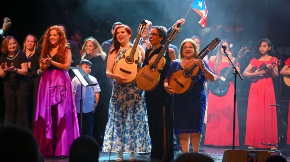 Cuatristas Maribel Delgado, Emma Colón Zayas, and Fabiola Méndez hold up their cuatros to the audience at the 25th Annual National Cuatro Festival as someone waves a Puerto Rican Flag in the back. 