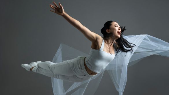 A dancer midair spread her hands open. She wears white pants and shit holding a white sheer piece of fabric. 