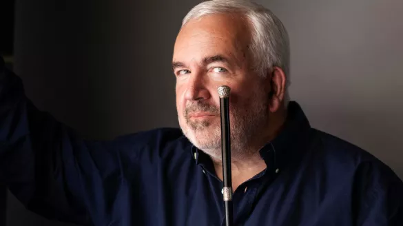 A gray-haired conductor holds a silver-tipped conducting baton to the corner of his mouth. 