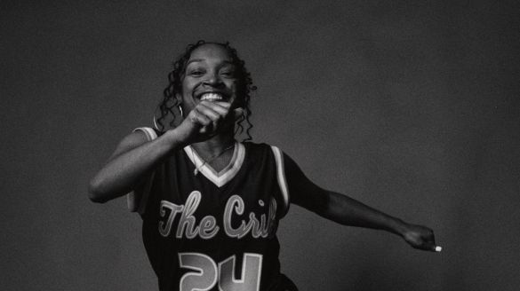 Black and white image of Donetta “Lilbit” Jackson dancing. Donetta smiles widely at the camera as she dances with her hands shuffling from the front to the side of her body. She wears a jersey, athletic shorts, and sneakers. The background is a deep grey making the darkness of her outfit stand out.