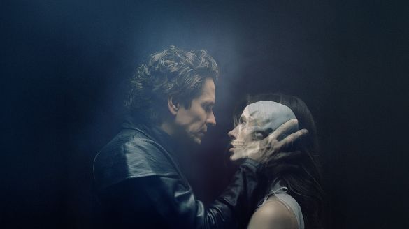 A man wearing black leather jacket holds someone whose face blurs between a womans and a skull.