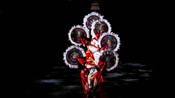 Dancers stand in a row wearing circular head pieces and bowing down.