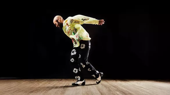 Jumaane Taylor dancing on a wooden floor. He wears black floral pants and a yellow printed long sleeve.