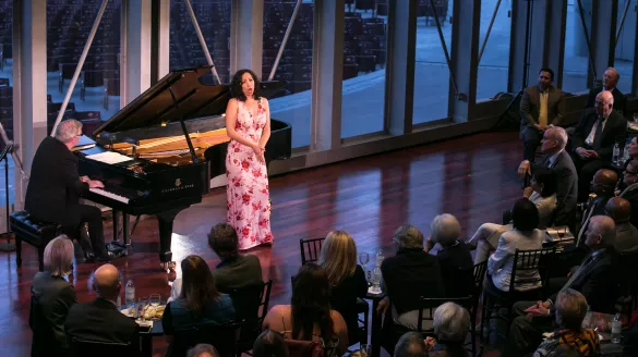 A woman wears a pink floral dress. She is singing in front of a seated audience and accompanied by a pianist.