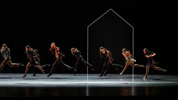 A group of dancer's mid performance jumping in the air. Only the dancers and an outline of a house is lit.