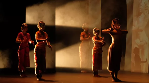 Five dancers stand wearing traditional Hindu attire. The stage is semi dark with the light coming through the back of the stage and large panels.