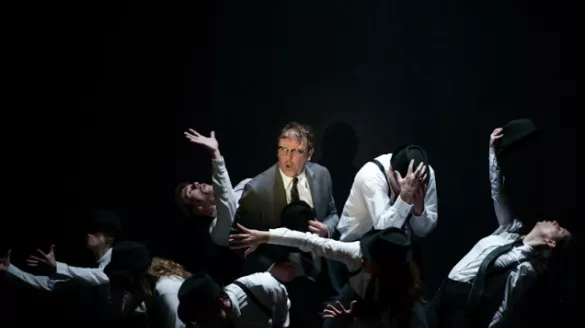 A group of dancers in black fedora hats and business suits gather around a man in a grey business suit with glasses.