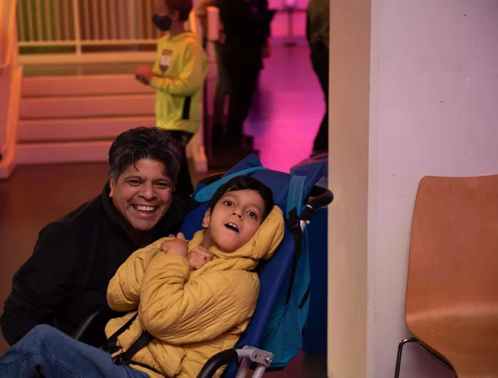 Father wears a black coat and son wears a yellow jacket, sitting in a blue wheelchair, smiling for the camera.