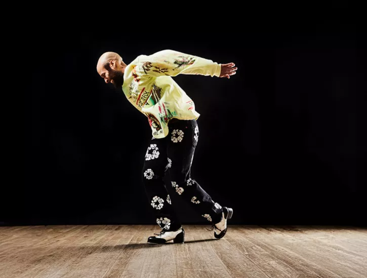 Jumaane Taylor dancing on a wooden floor. He wears black floral pants and a yellow printed long sleeve.